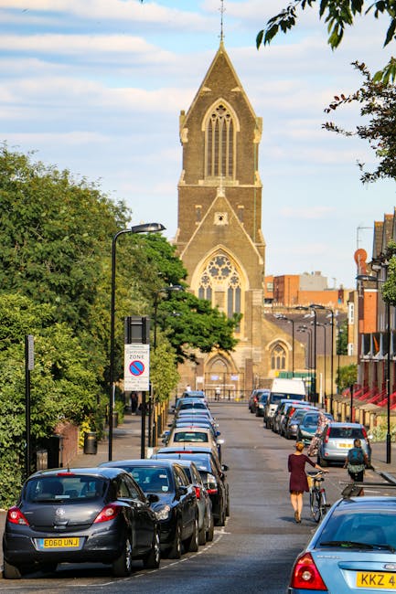 A narrow urban street scene in Dulwich featuring a row of parked cars on both sides, including compact and hatchback models, with some vehicles partially shaded by nearby leafy trees. Two pedestrians are visible walking along the pavement; one person in a crimson coat pushing a bicycle and another in casual attire. In the background, the prominent Gothic-style church with a tall, pointed spire, arched windows, and detailed stonework dominates the scene. The street is equipped with standard black lampposts and street signage indicating parking restrictions. Building facades along the street include brick and modern materials, with some featuring shop fronts or residential entrances. Overhead, the sky is partly cloudy, providing natural lighting that highlights the scene's details. This image relates to house removals and relocation services, illustrating the typical environment where Man with Van Dulwich operates, including loading and logistics processes involved in furniture transport and home relocation in the Dulwich SE22 area.