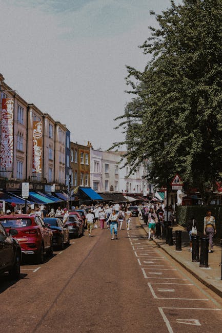 A busy street scene on Lordship Lane in Dulwich SE22 with parked cars lining both sides of a narrow roadway, which is designated for parking and loading with clearly marked bays. Pedestrians, including individuals and small groups, are walking along the pavement and crossing the street. On the left side, a row of multi-storey shops with colorful facades and awnings extends along the street, providing retail and service outlets that are typical of a vibrant local shopping area. Large trees with green foliage shade parts of the sidewalk, while street signs and bollards help organise traffic and pedestrian flow. In the background, a mixture of residential and commercial buildings rises beyond the shops, indicating an active community environment. The scene captures the typical urban atmosphere where home relocation and furniture transport activities might take place, with potential for vehicles from companies like Man with Van Dulwich to load furniture and boxes, utilizing nearby parking and access points during an ongoing moving or packing process.