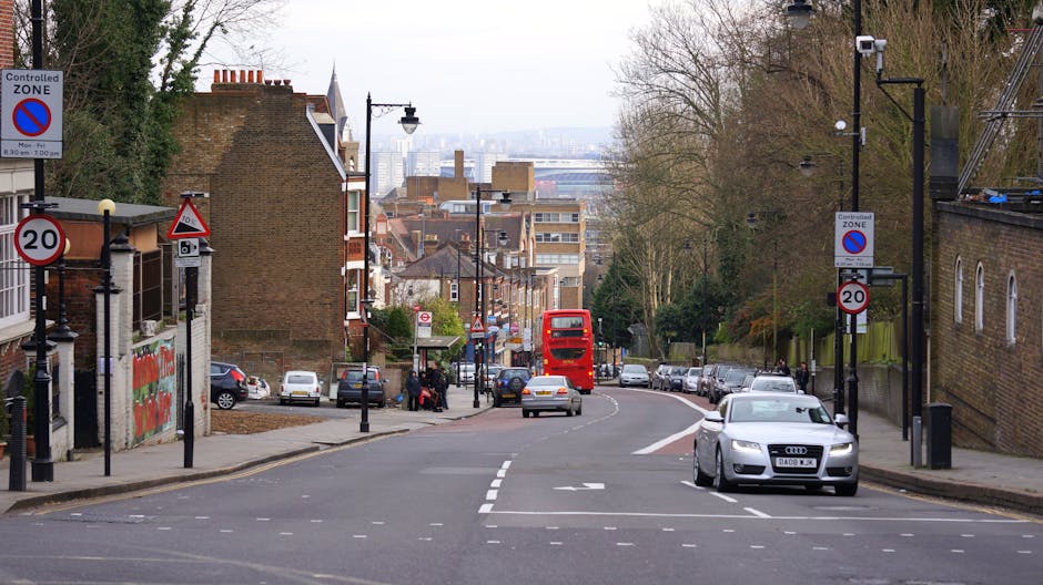 A narrow urban street scene in Dulwich featuring a row of parked cars on both sides, including compact and hatchback models, with some vehicles partially shaded by nearby leafy trees. Two pedestrians are visible walking along the pavement; one person in a crimson coat pushing a bicycle and another in casual attire. In the background, the prominent Gothic-style church with a tall, pointed spire, arched windows, and detailed stonework dominates the scene. The street is equipped with standard black lampposts and street signage indicating parking restrictions. Building facades along the street include brick and modern materials, with some featuring shop fronts or residential entrances. Overhead, the sky is partly cloudy, providing natural lighting that highlights the scene's details. This image relates to house removals and relocation services, illustrating the typical environment where Man with Van Dulwich operates, including loading and logistics processes involved in furniture transport and home relocation in the Dulwich SE22 area.