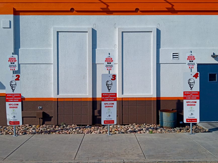 The image shows the exterior wall of a commercial building with a light grey painted surface and a brown lower section, located at East Dulwich station. In front of the wall, on the sidewalk, there are three vertical signs supporting a house removals service, each marked with bold red numbers 2, 3, and 4, indicating different pickup points. The signs display the KFC logo and advertise quick pickup for mobile orders with the website links KFC.COM and GRUBHUB, along with instructions for home delivery drivers. The sidewalk is made of concrete paving slabs, with a strip of small, multicoloured stones along the building’s base. To the right, there is a metal barrel, potentially used for disposal or storage. The scene suggests an area designated for logistical organisation in a home relocation process, with the signage preparing for furniture transport or moving tasks, possibly related to the services offered by Man with Van Dulwich.