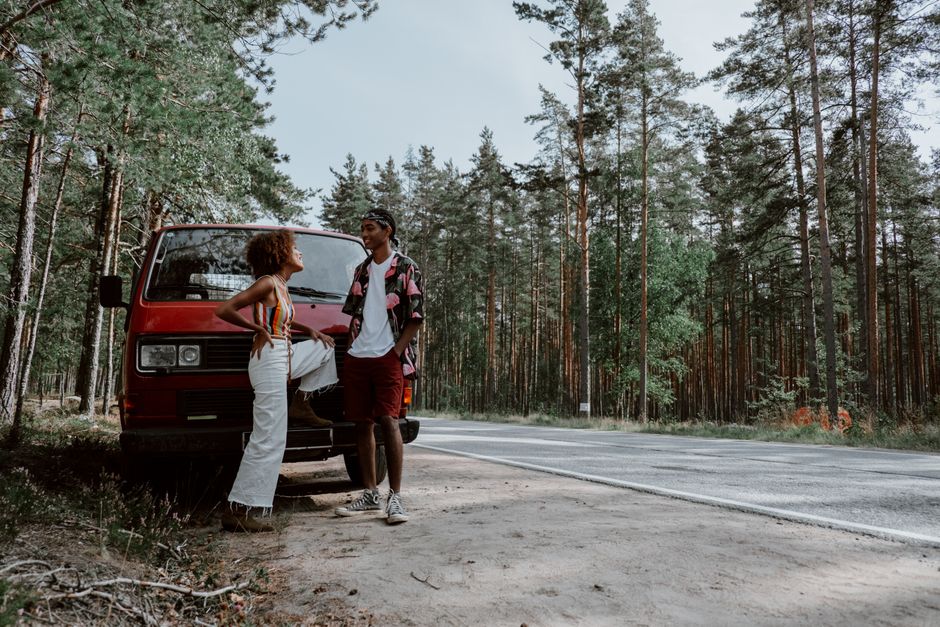 A man and a woman stand beside a red van parked on the side of a rural road surrounded by tall pine trees during daytime. The woman is wearing a sleeveless striped top and white pants, while the man is dressed in a short-sleeved shirt with a floral pattern and dark shorts. Both are smiling and engaging in conversation, with the woman placing her hand on her hip and the man standing with his hands in his pockets. The van appears to be used for home relocation, as it is positioned close to the pavement with the rear doors possibly open for loading or unloading furniture and boxes. Inside or near the van, there are visible packages wrapped in plastic, cardboard boxes, and furniture pieces, indicating preparations for a moving or packing process. The natural lighting highlights the outdoor setting, emphasizing the transport and logistics aspect typical of house removal services, with the presence of trolleys or straps not visible in this image.